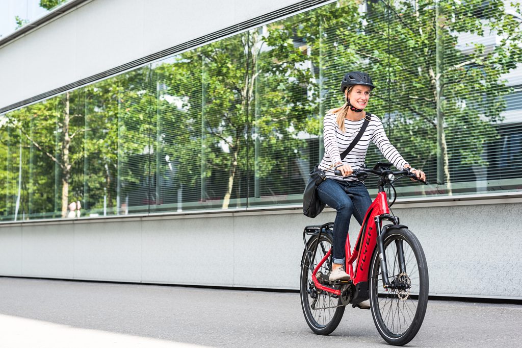 junge Frau auf E-Bike mit Helm und Tasche im urbanen Setting, könnte am Weg in die Arbeit sein