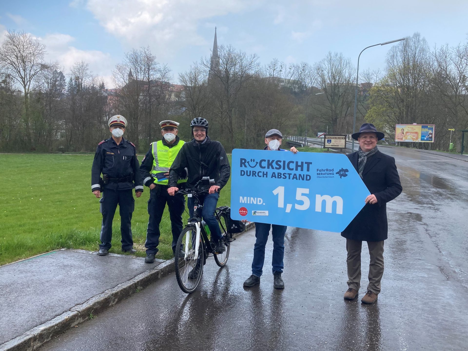 zwei Polizisten, ein Radfahrer, zwei Männer, die ein Schild mit der Aufschrift "Rücksicht durch Abstand, mind. 1,5m" halten