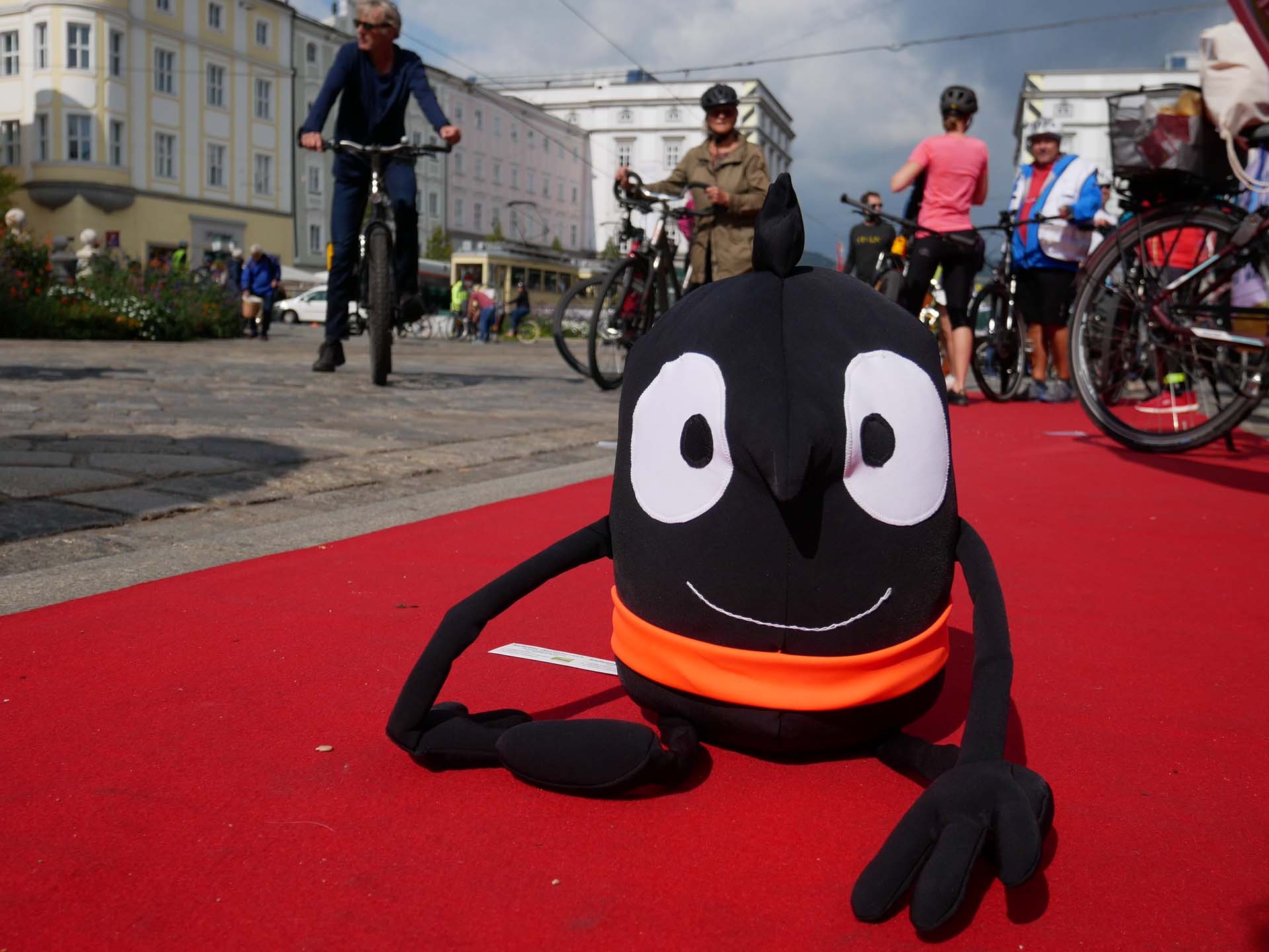 Das Maskottchen der Mobilitätswoche Edgar sitzt auf einem roten Teppich. Im Hintergrund ist der Stadtplatz Linz und mehrere Radfahrer sichtbar.