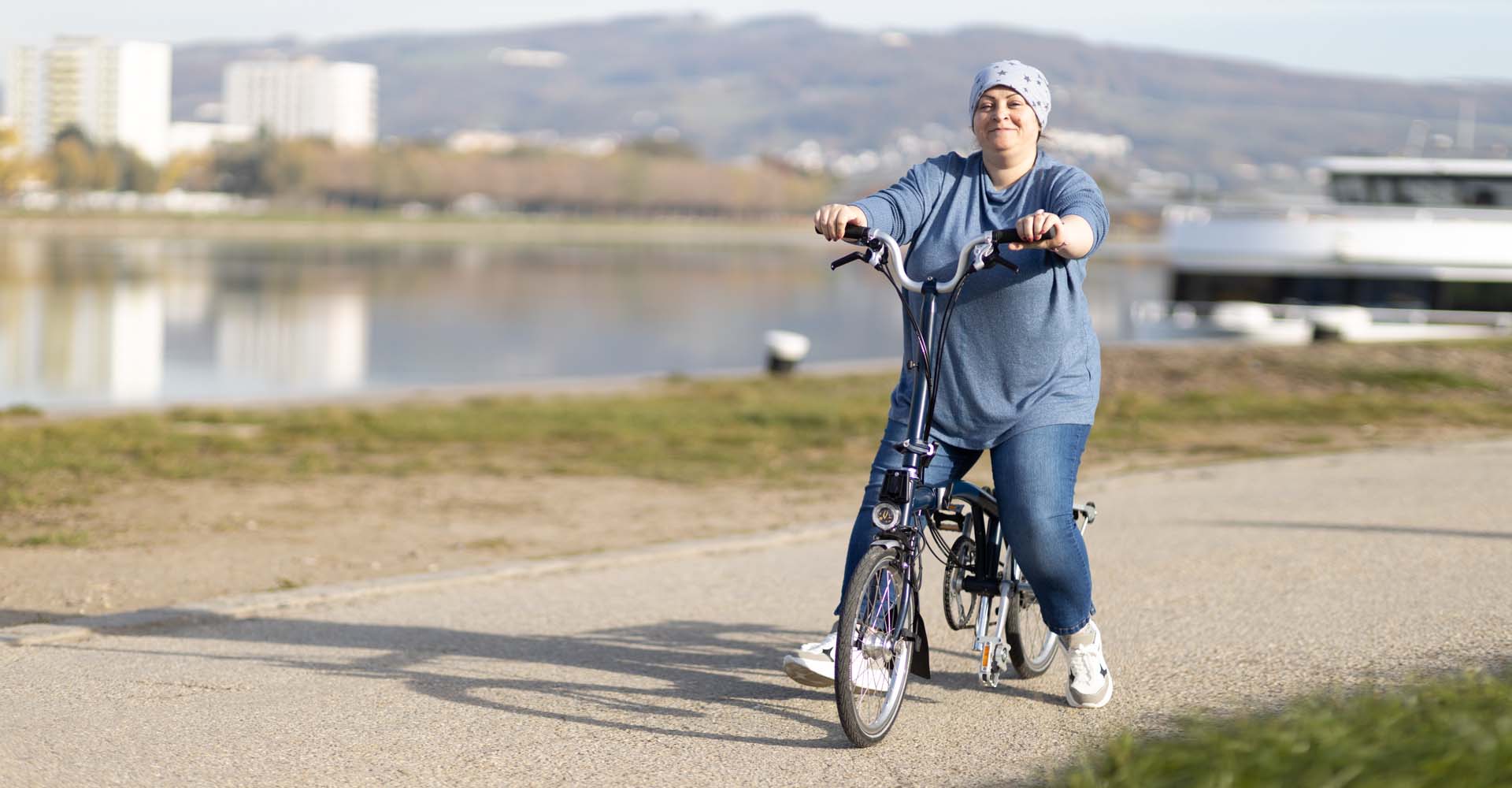 Eine lächelnde Radfahrerin mit Klapprad fährt auf einem Weg neben einem Fluss.