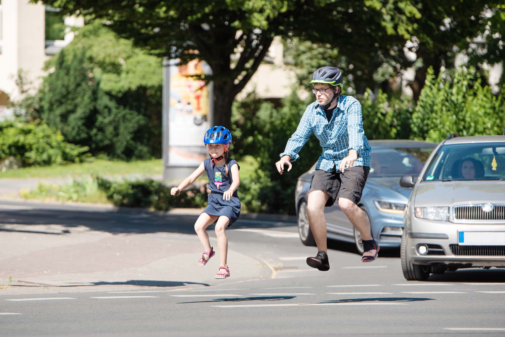 Ein Mädchen und ein Herr radeln mit unsichtbaren Rädern. 2 Autos warten im Hintergrund.