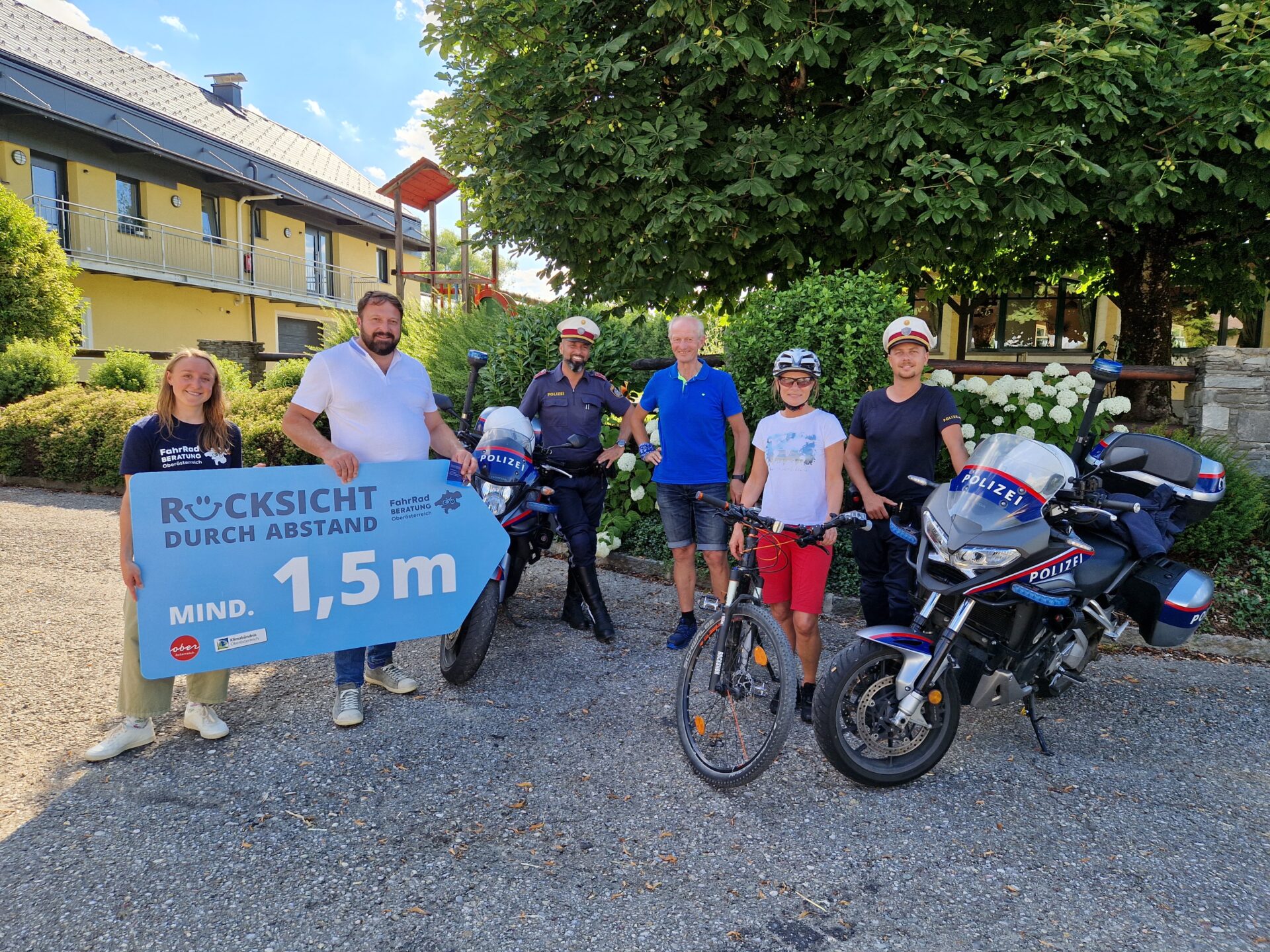 4 Personen und 2 Polizisten posieren mit Fahrrad, Polizei-Motorräder und einem Plakat mit der Aufschrift "Rücksicht durch Abstand - mind. 1,5m