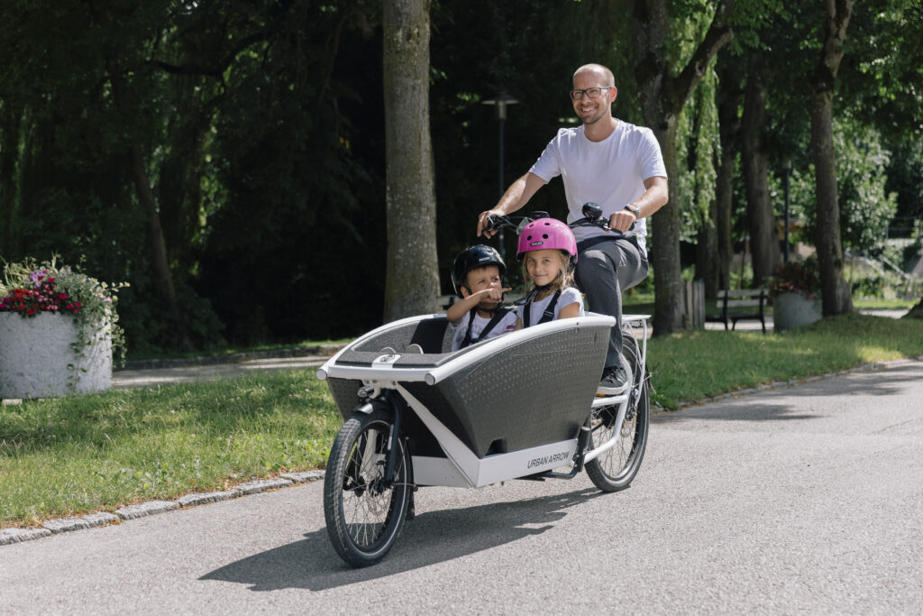 Lachender Mann mit Urban Arrow Cargobike, zwei mitfahrende Kinder