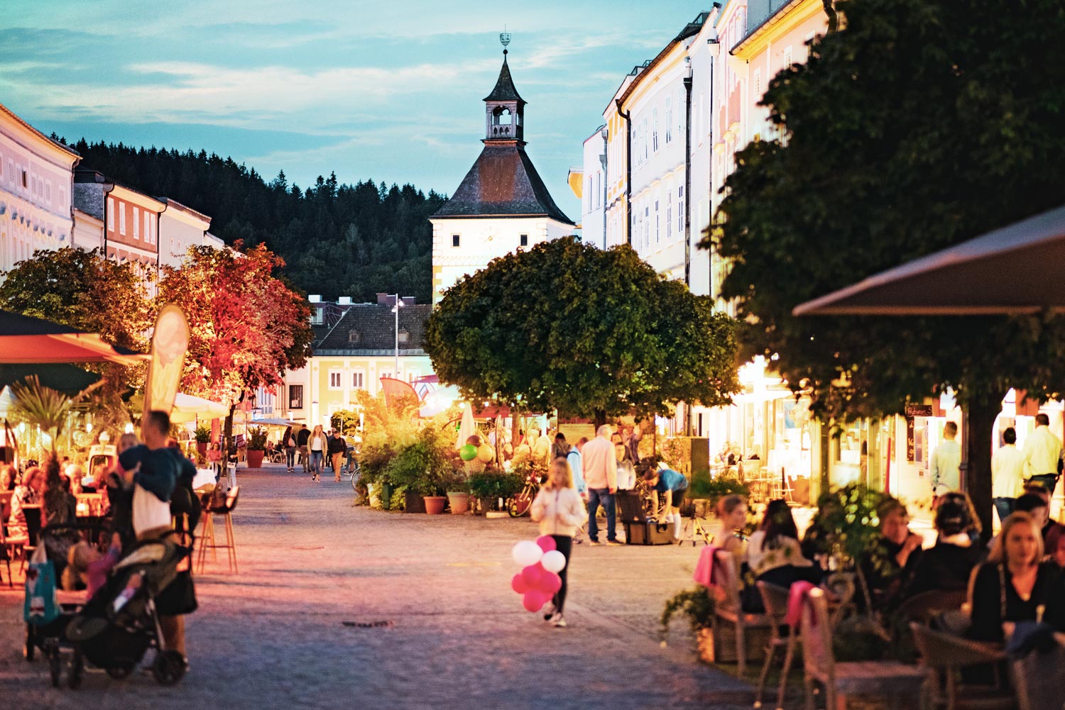 Abendliche Aufnahme im Stadtplatz Vöcklabruck. Personen sitzen im Außenbereich der Gastronomiebetriebe. Ein Mädchen spielt mit Luftballons.