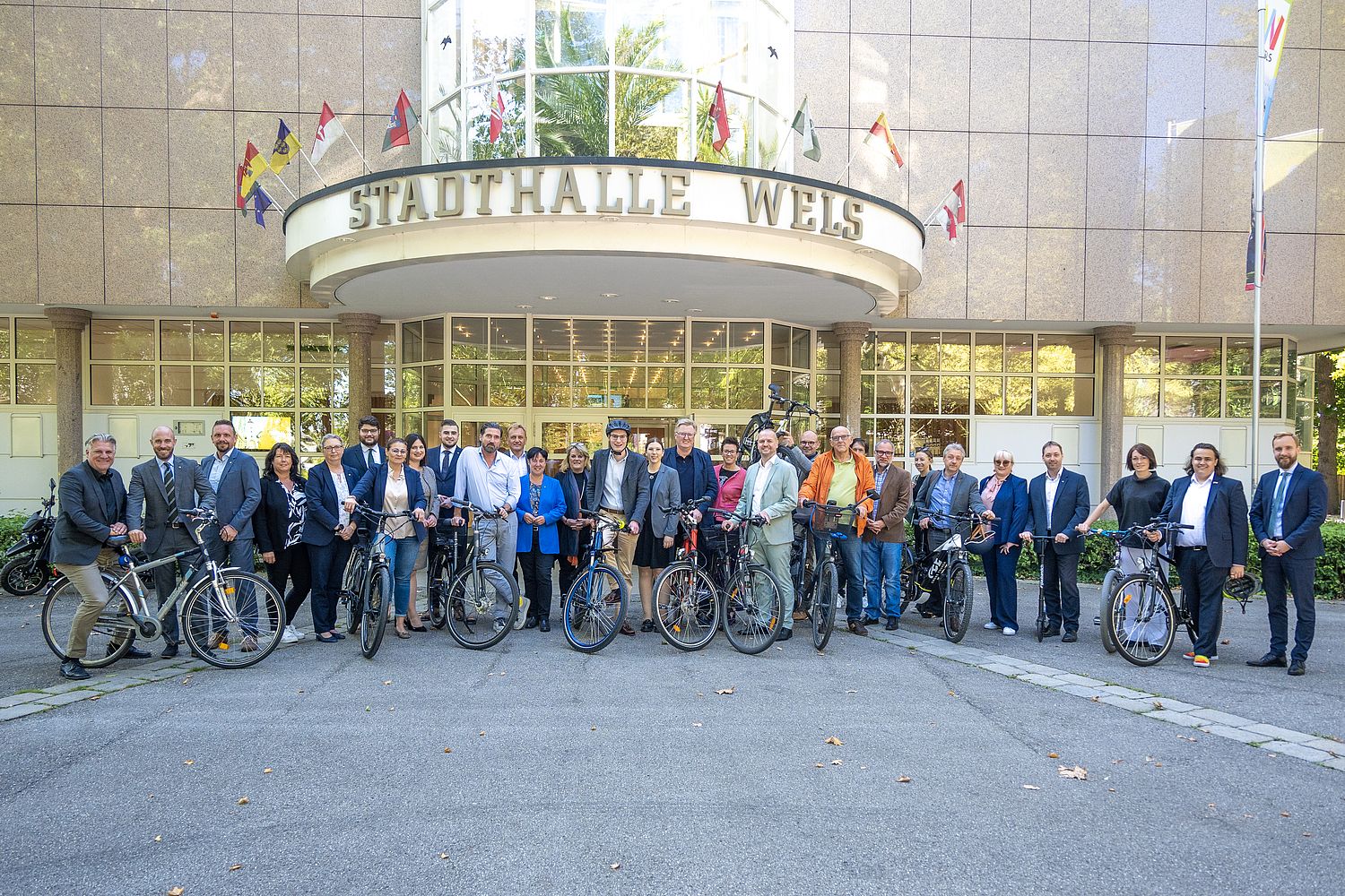 Gruppenfoto mit vielen Personen in Anzügen vor der Stadthalle Wels, einige haben ein Fahrrad