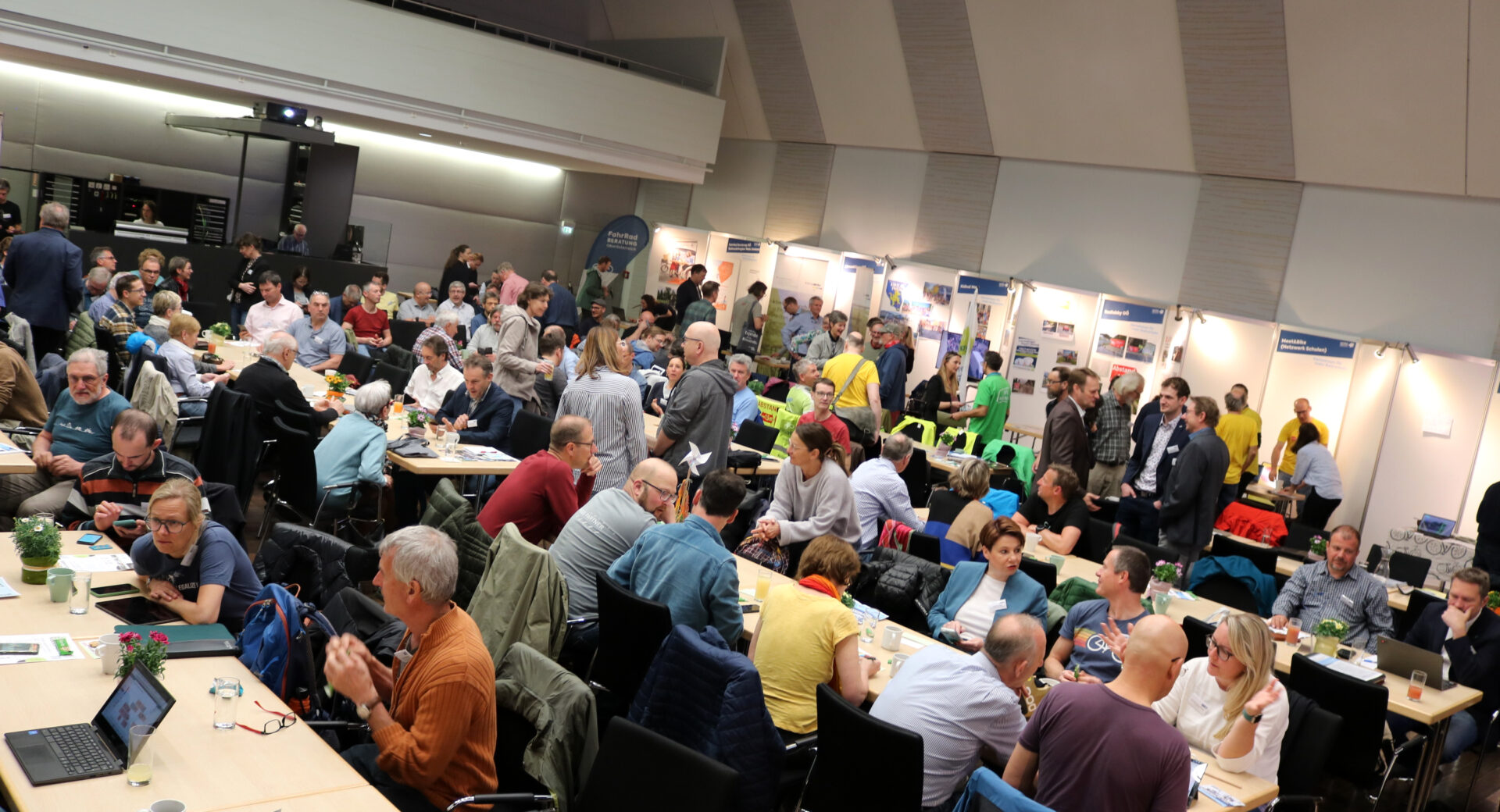 Saal2, sehr viele Menschen in einem Saal, manche sitzen an Tischen