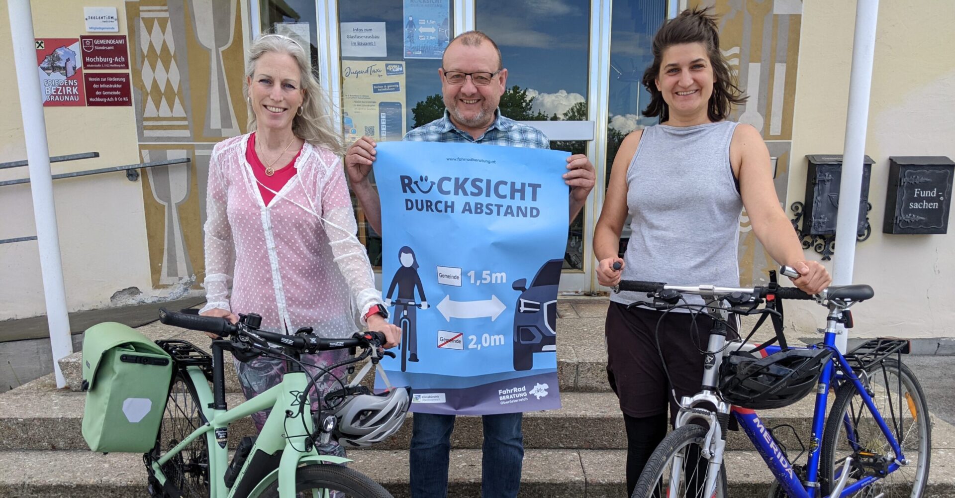 drei Personen, zwei von ihnen mit Fahrrädern, ein Mann hält ein "Rücksicht mit Abstand" - Plakat