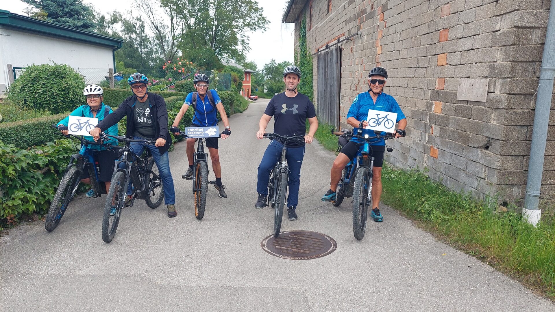 fünf Personen auf ihren Fahrrädern auf einer schmalen Straße, drei von ihnen halten ein Schild von der Fahrradberatung