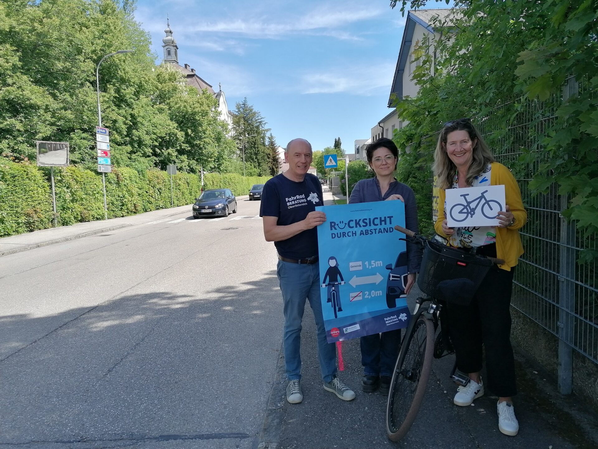 drei Personen mit einem Fahrrad am Rand einer Straße in einem Dorf, sie halten ein "Rücksicht durch Abstand" - Plakat