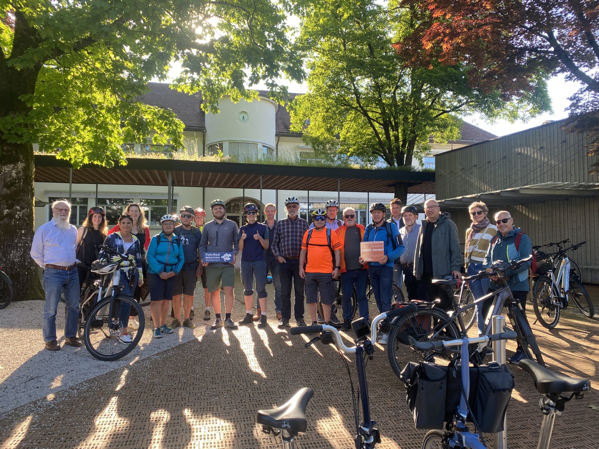 viele Radfahrerinnen und Radfahrer nebeneinander stehend unter Bäumen, im Vordergrund die Fahrräder