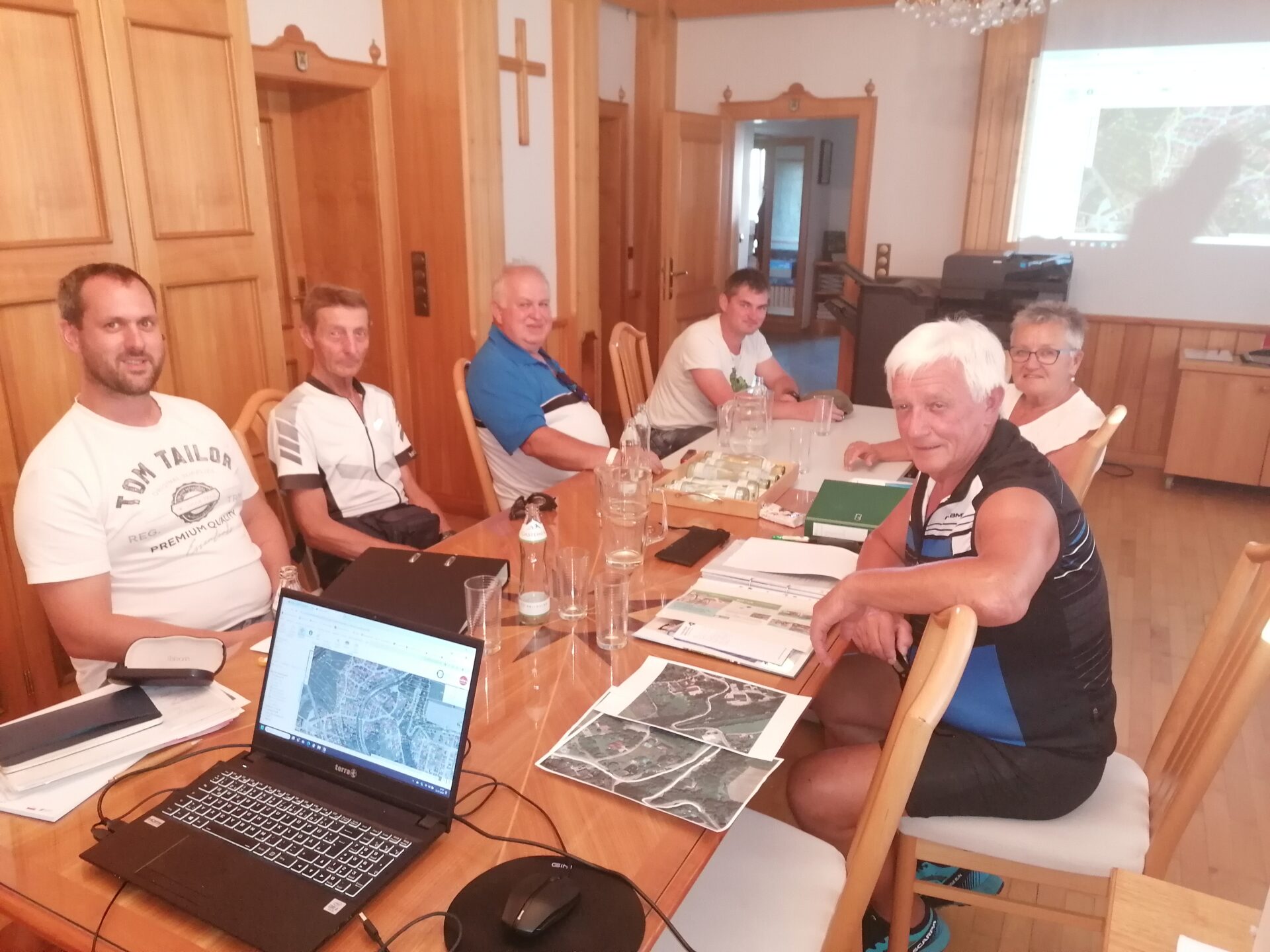 sechs Personen sitzen an einem Tisch in einem Innenraum, am Tisch sind verschieden Karten und ein Laptop