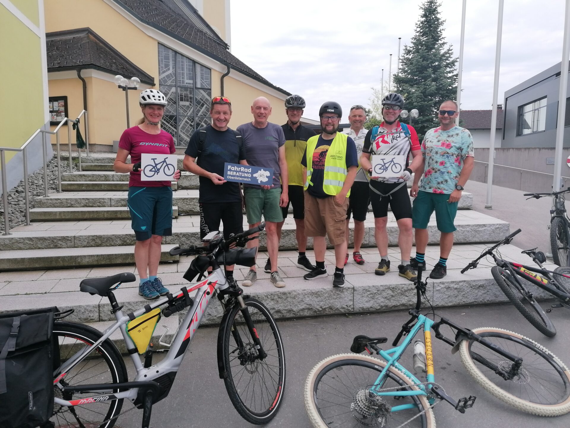 Personen, in die Kamera schauend, mit "Fahrradberatung"s- Schildern in der Hand, vor ihnen die Fahrräder