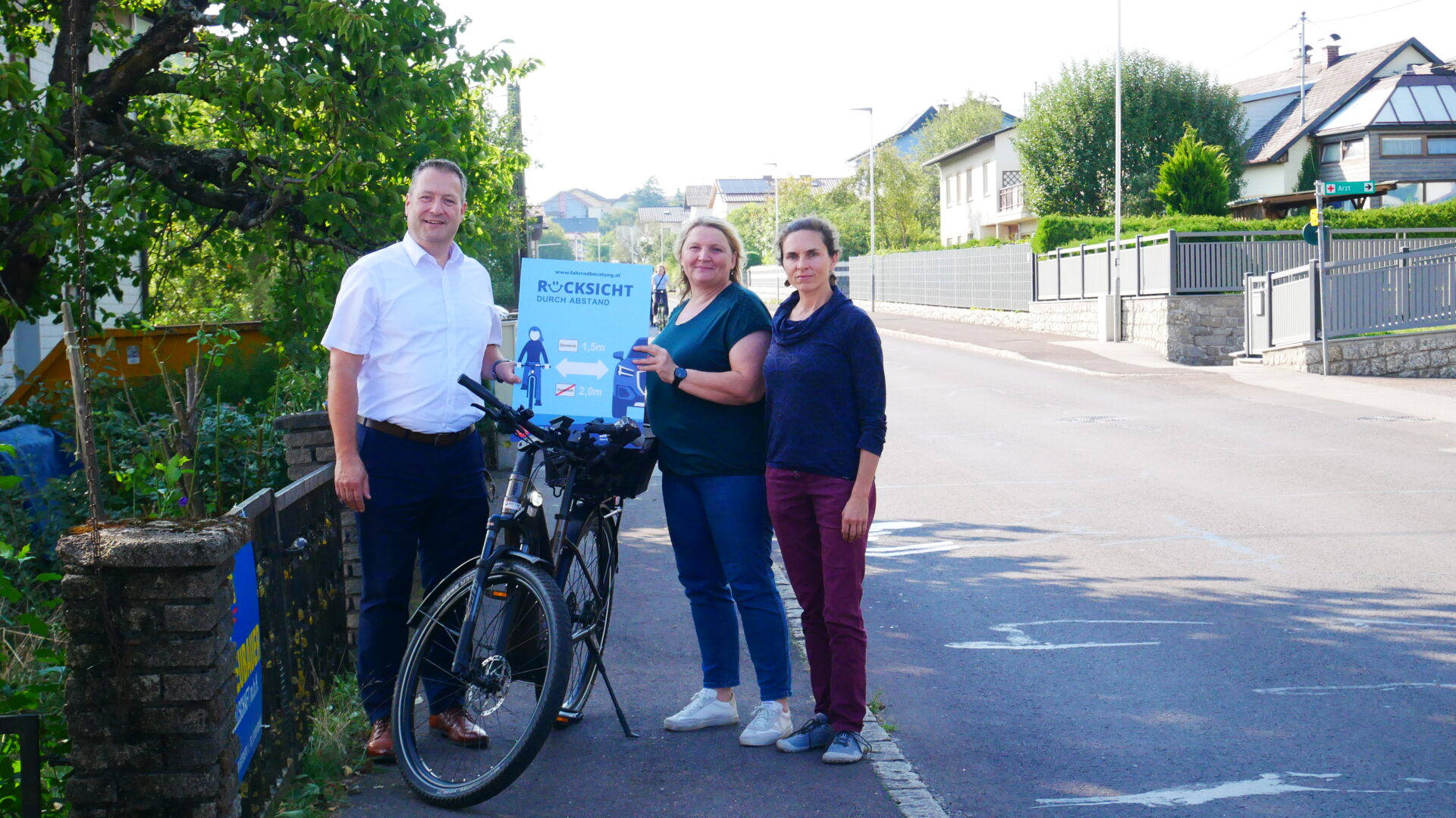 Drei Menschen mit einem Fahrrad halten ein Schild mit der Aufschrift "Rücksicht durch Abstand"