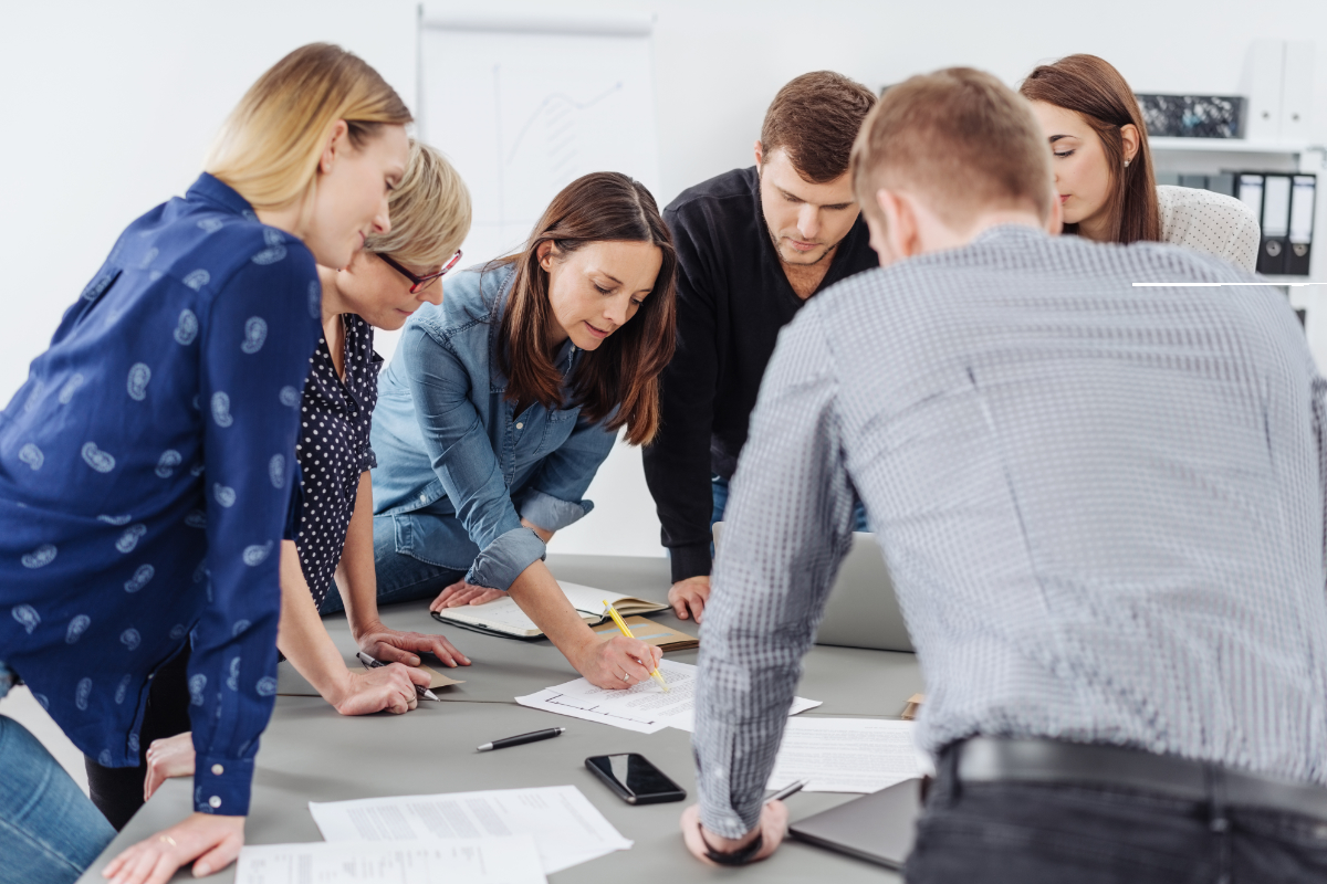 Motivated group of young business people grouped around a table listening to a female team leader or manageress explain paperwork