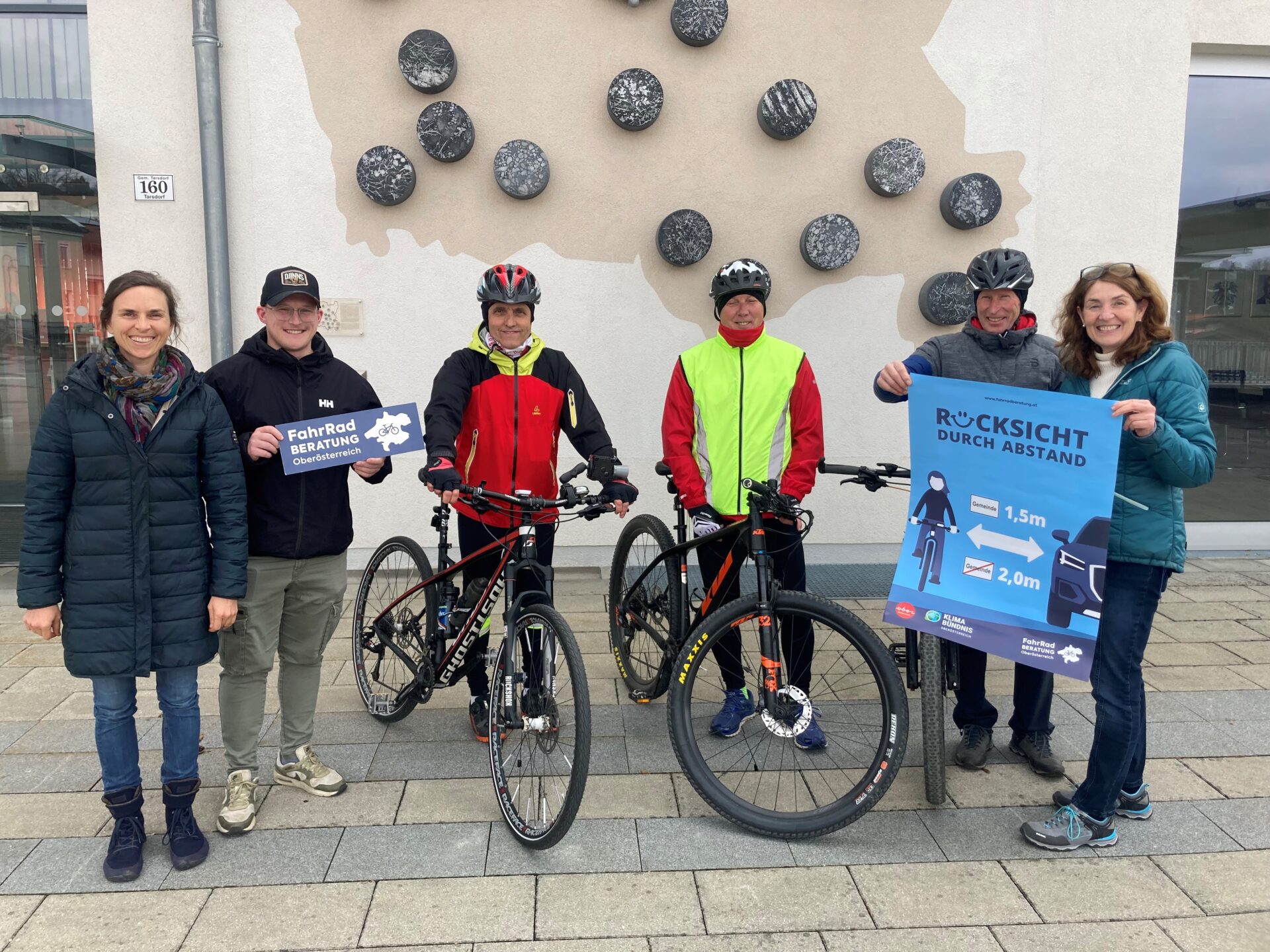 Gruppe von Menschen tlw. mit Fahrrädern und Helmen vor Gemeindeamt in Tarsdorf, halten ein Plakat "Rücksicht durch Abstand"