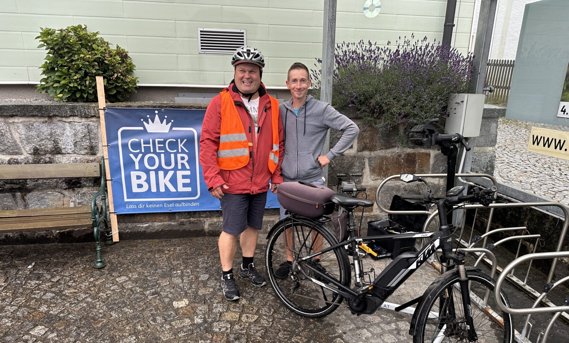 Zwei Personen stehen neben einem Fahrrad vor einem „Check Your Bike“-Schild.