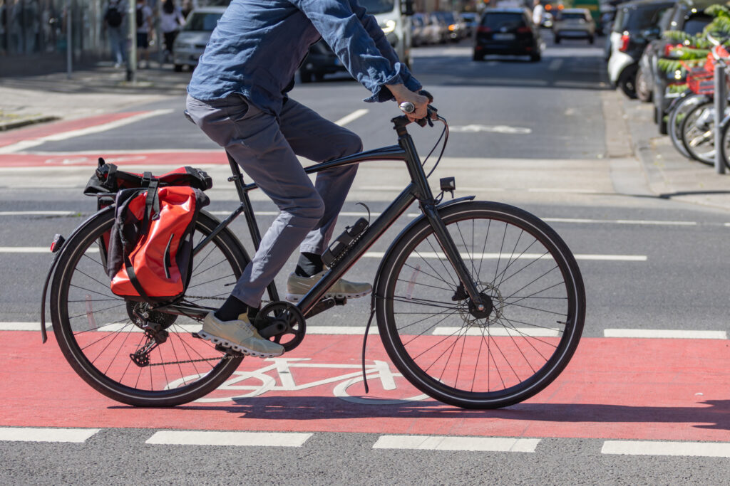 Fahrrad fährt über Kreuzung mit roter, flächiger Markierung, Kopf abgeschnitten. Fahrradtaschen am Gepäckträger