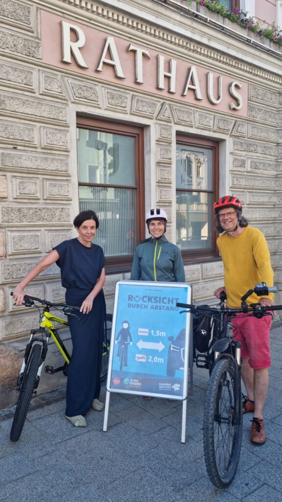 3 Personen vor Rathaus mit 2 Fahrrädern, mit Plakatständer, darin "Rücksicht durch Abstand" Plakat